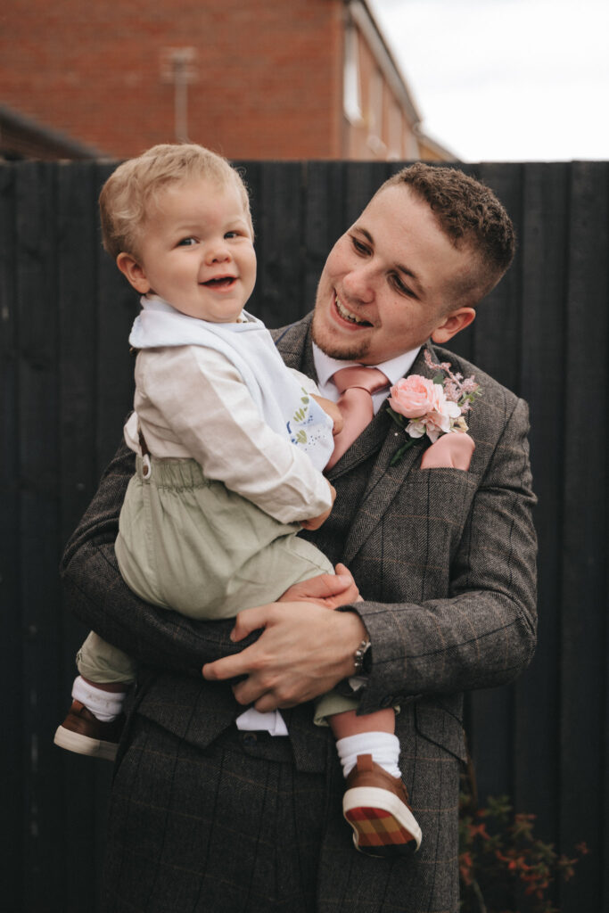 A smiling man in a gray patterned suit, perfect for wedding photography, holds a happy toddler in his arms. The toddler, wearing green overalls and a white shirt, smiles at the camera. The man, with a pink flower boutonniere, looks fondly at the child as they stand by a dark fence and brick building. © Aimee Lince Photography