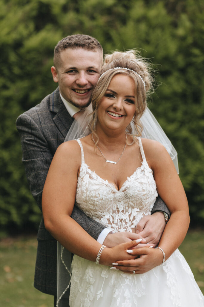 A couple stands together outdoors at the picturesque Oaklands Hall Hotel, embraced by lush greenery. The man, in a gray suit and tie, wraps his arms around the woman in a white lace wedding dress and veil. Their smiles radiate pure joy, perfectly captured by wedding photography for this special occasion. © Aimee Lince Photography