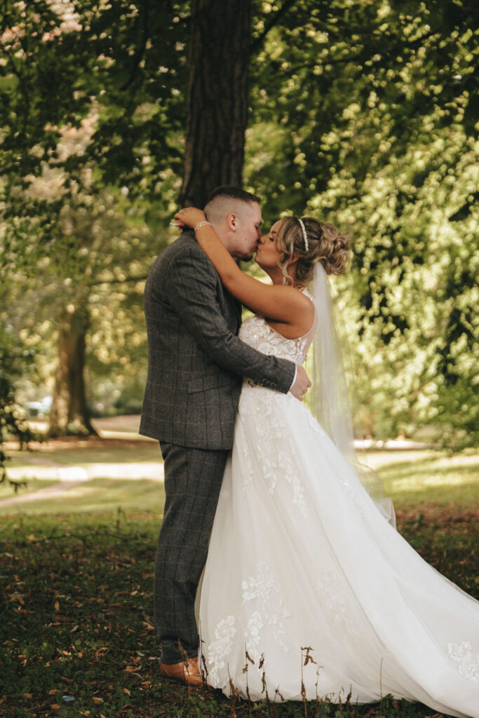 Under the sprawling canopy of a large tree in a sunlit park, a couple shares a tender kiss. The groom, clad in a gray suit, and the bride, adorned in a white lace wedding dress with veil, are framed by lush green foliage at Oaklands Hall Hotel, creating an utterly romantic atmosphere. © Aimee Lince Photography