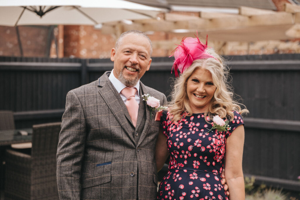 A smiling man and woman pose together outdoors, perfect for wedding photos. The man dons a gray plaid suit with a pink tie and boutonniere, while the woman stuns in a navy dress with pink floral patterns and a matching fascinator, set against the dark fence of Oaklands Hall. Patio chairs and an umbrella stand behind them. © Aimee Lince Photography