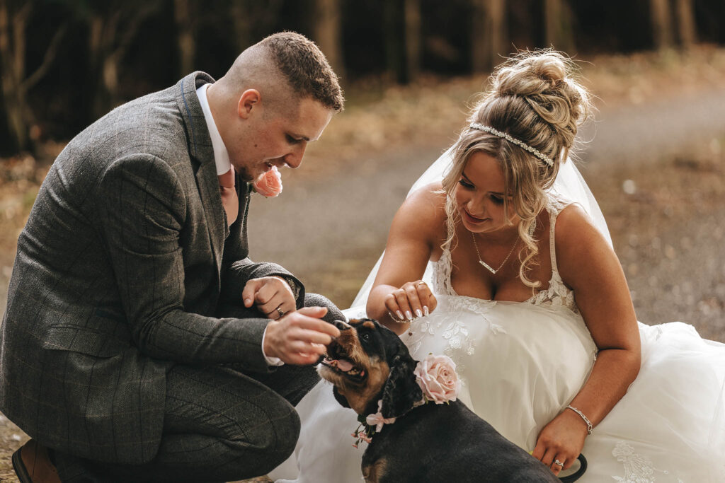 A bride and groom kneel on a forest path, smiling as they pet a small black and brown dog adorned with pink flowers. Captured by the talented team at Oaklands Hall, the scene radiates joy and intimacy, with soft, natural lighting filtering through the trees—a perfect moment of wedding photography. © Aimee Lince Photography