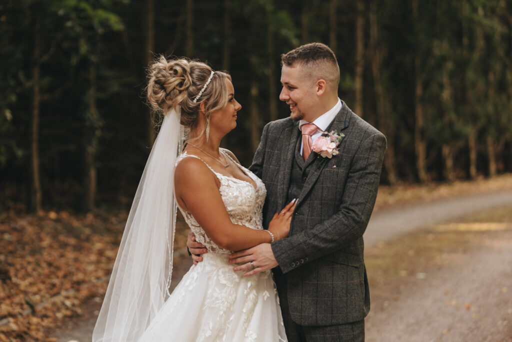 The bride and groom gaze at each other lovingly, captured in stunning wedding photography. The bride wears a white lace gown and veil, her hair styled up. The groom is in a gray checkered suit with a pink tie. They stand on a path surrounded by trees with autumn leaves scattered on the ground. © Aimee Lince Photography