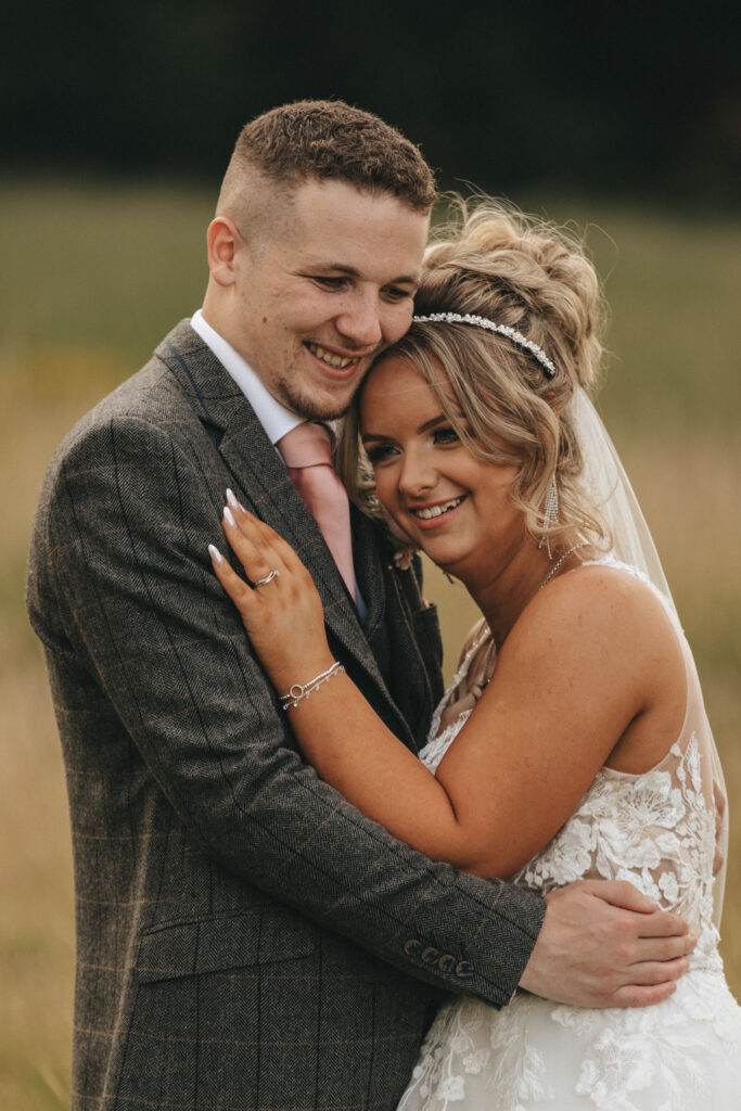 A bride and groom embrace outdoors at The Oaklands. The groom wears a gray plaid suit with a pink tie, while the bride dons a lace wedding dress and pearl headband. Her hair is styled in an updo, and they both smile warmly, captured beautifully in this romantic wedding photography amidst a blurred grassy field. © Aimee Lince Photography