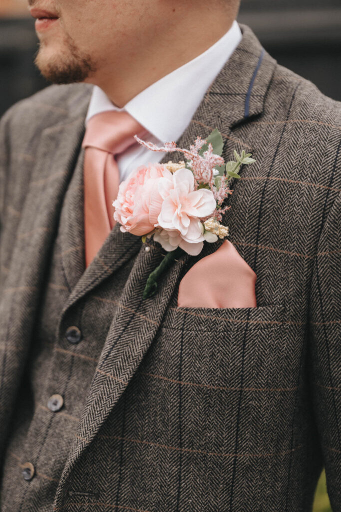Close-up of a person in a tweed suit with a white shirt at Oaklands Hall. They sport a peach-colored tie and matching pocket square, perfect for wedding photography. A boutonniere with pink and white flowers adorns the lapel, complementing the subtle plaid pattern as they face left, slightly showing a beard. © Aimee Lince Photography