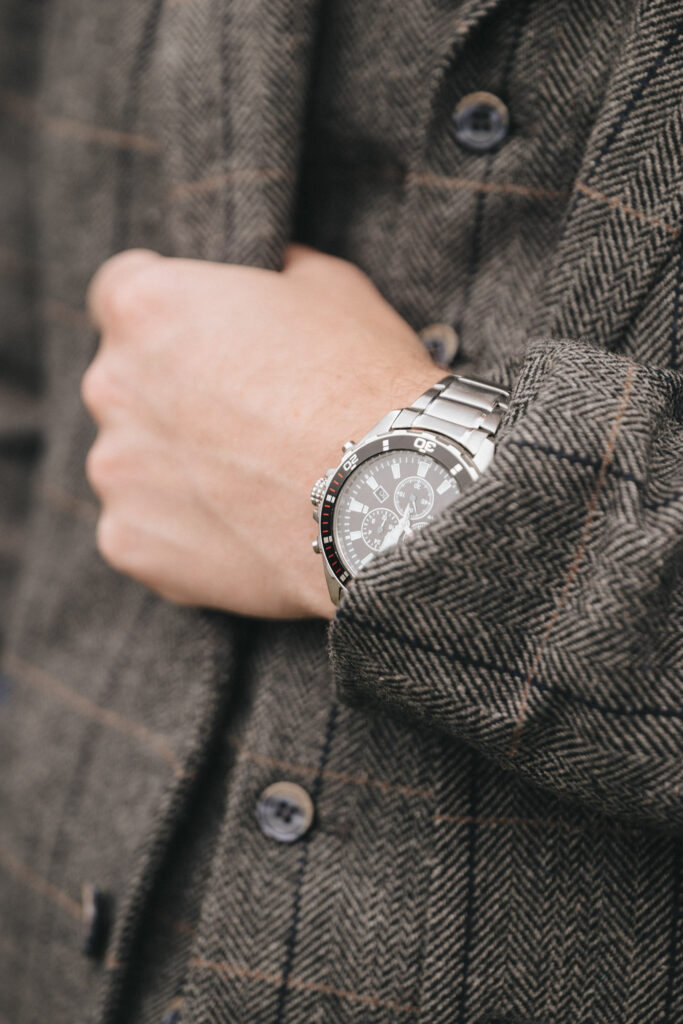 Close-up of a person at Oaklands Hall, wearing a herringbone-patterned tweed jacket with dark buttons. They adjust the jacket with their left hand, revealing a silver chronograph watch with a black dial and metal bracelet. The setting subtly enhances the elegance captured in wedding photos. © Aimee Lince Photography