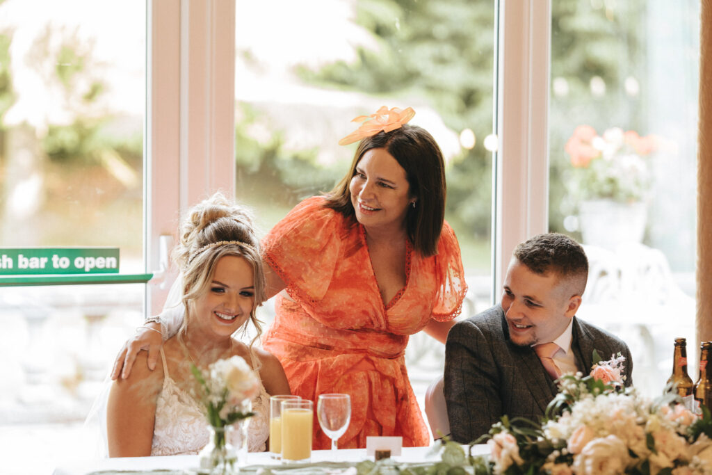 The Oaklands Hall Hotel Wedding Photographer - Loren & Bailey © Aimee Lince Photography (654) A woman in an orange dress with a hat stands smiling, placing her hand on the shoulder of a bride at the Oaklands Hall Hotel wedding. The bride is seated, wearing a white dress and veil. A man in a suit sits next to her, smiling. They are surrounded by flowers and drinks, with a glass door in the background. © Aimee Lince Photography