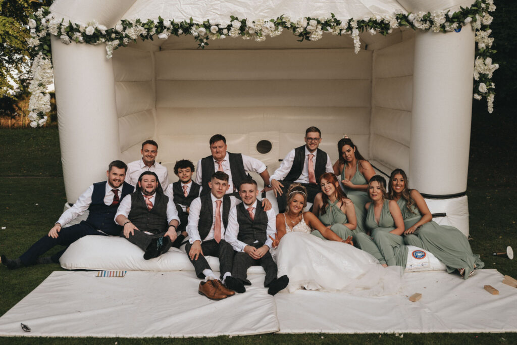 A group of people dressed in formal attire, including suits and long dresses, sit on a white inflatable structure decorated with flowers at what appears to be a wedding. Some are seated on the ground, all appearing joyful against the backdrop of Oaklands Hall’s verdant greenery. © Aimee Lince Photography