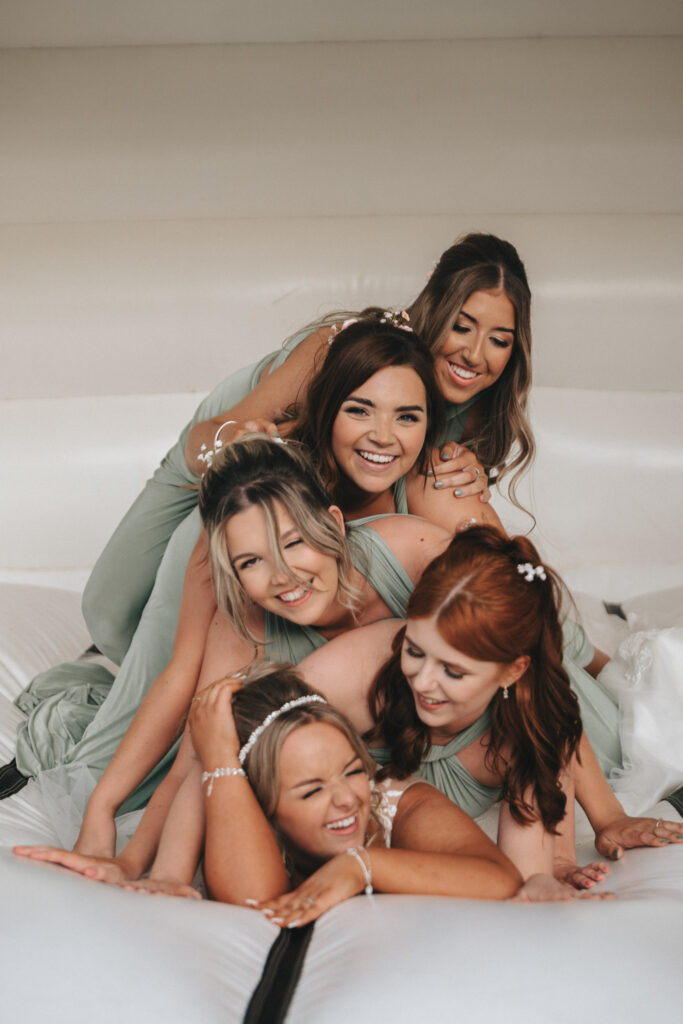 Five smiling women in pastel green dresses are playfully stacked on a white inflatable surface at Oaklands Hall Hotel. With various hairstyles and floral hair accessories, this joyful scene captures a wedding photo moment filled with carefree laughter and the magic of celebration. © Aimee Lince Photography
