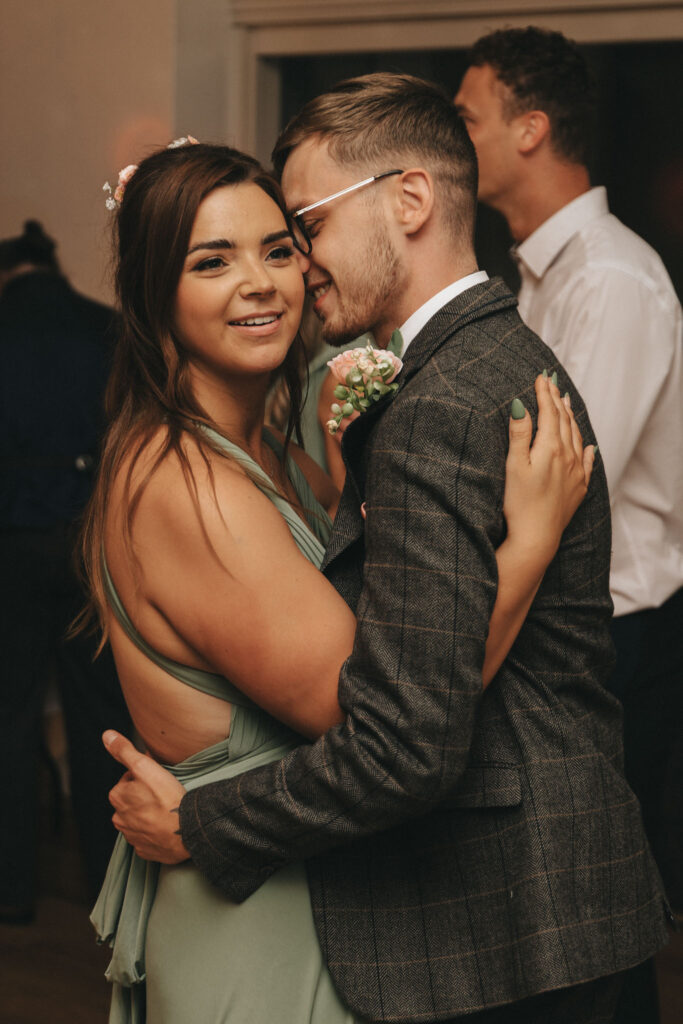 In a tender embrace, the couple dances closely. The woman dazzles in a sleeveless green dress, and the man wears a gray plaid suit adorned with a boutonniere. Both are beaming amidst a blurred backdrop of guests at Oaklands Hall Hotel—a perfect moment captured in wedding photography. © Aimee Lince Photography