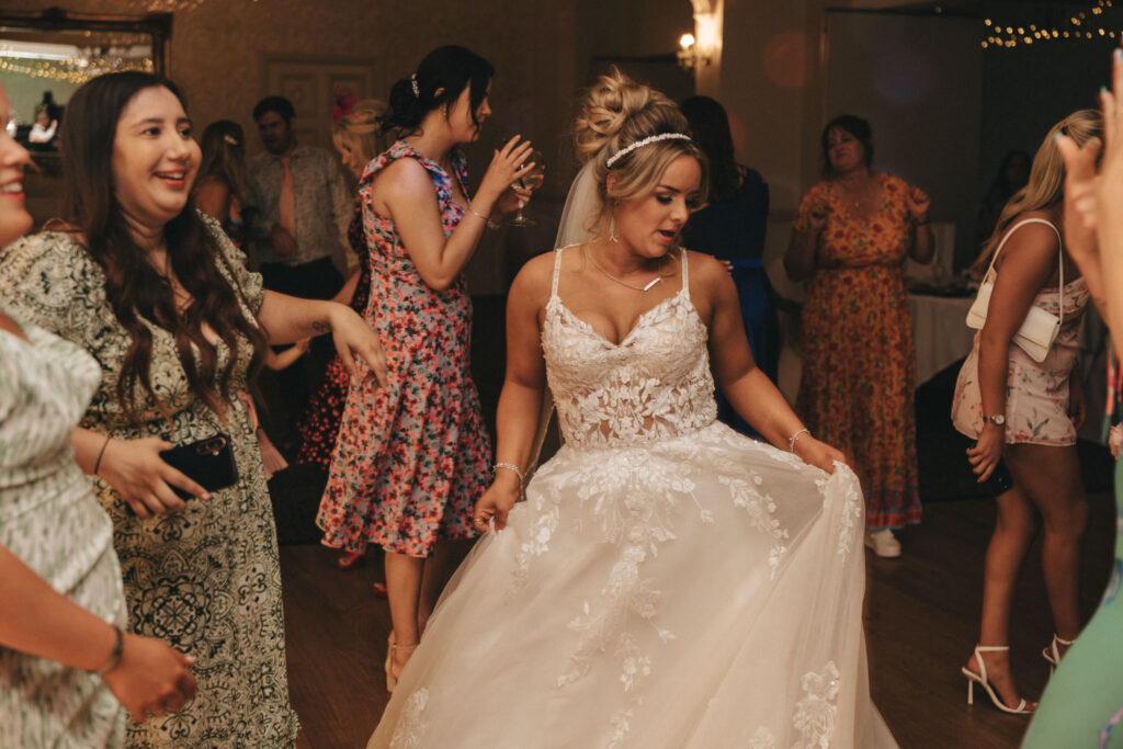 A bride in an elegant lace wedding dress dances at a reception with several guests at the charming Oaklands Hall Hotel. She holds her skirt while people around her, dressed in various floral and patterned outfits, enjoy the celebration. Warm lighting highlights decorations captured in stunning wedding photography. © Aimee Lince Photography