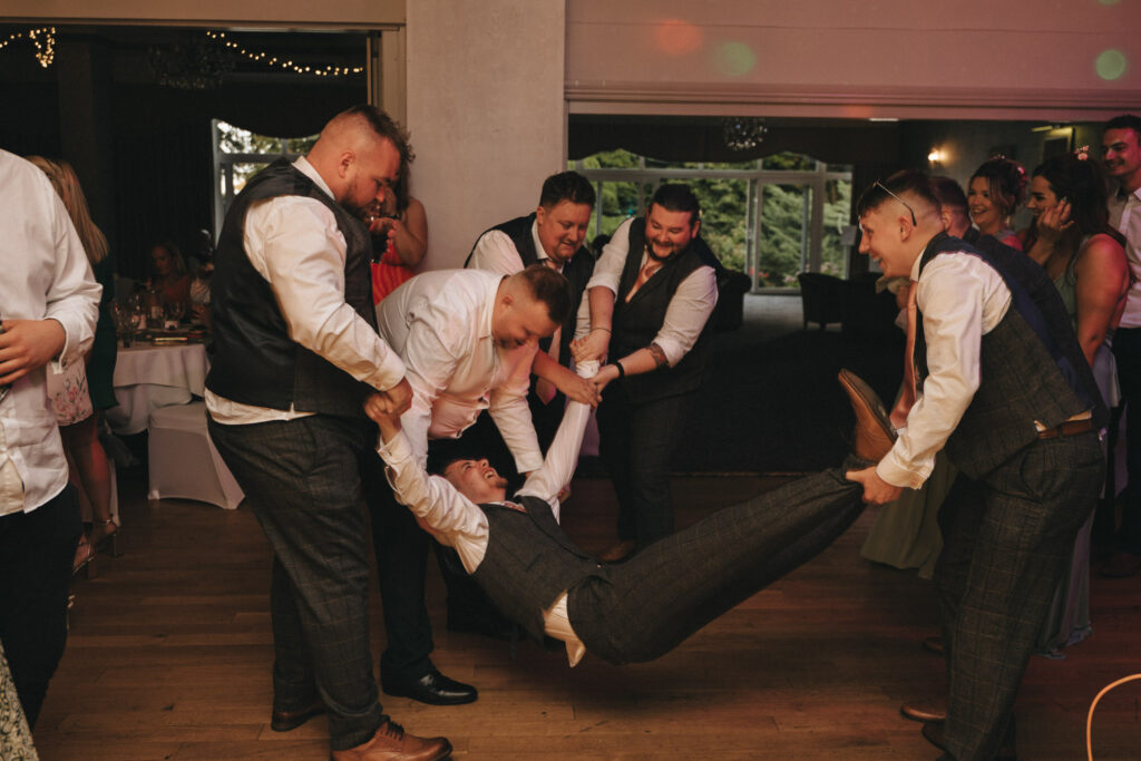 A joyful scene unfolds at the wedding reception in Oaklands Hall, where a group of well-dressed men playfully lift another smiling man in a suit. Indoors, large windows reveal lush greenery and festive lights twinkle above as guests mingle, all captured beautifully through wedding photography. © Aimee Lince Photography