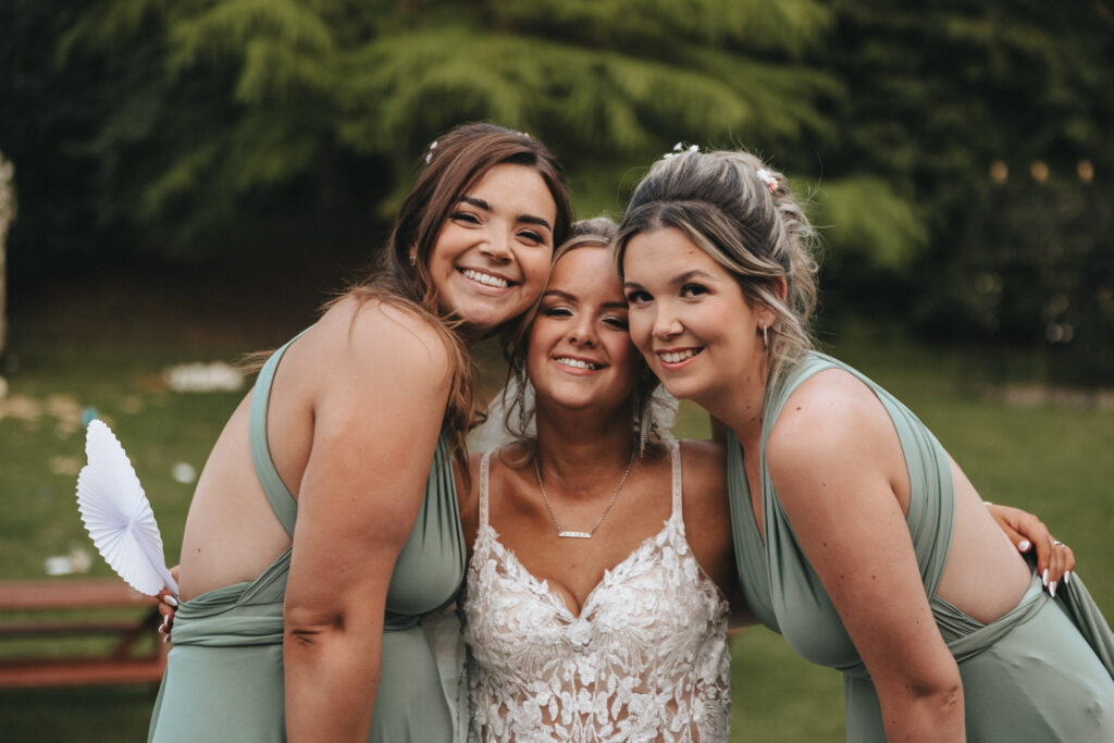 Three women beam outdoors at what appears to be a wedding, possibly at Oaklands Hall Hotel. The bride in the center is resplendent in a white lace dress, flanked by bridesmaids in sage green. Trees and a blurred background with a picnic table are visible as one bridesmaid holds a white fan. © Aimee Lince Photography