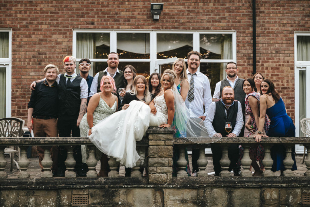 A group of people stands on a stone terrace at the Oaklands Hall Hotel in front of a brick building with large windows. Dressed in formal and semi-formal attire, they exude a celebratory wedding atmosphere. Most are smiling, and string lights twinkle through the windows. © Aimee Lince Photography