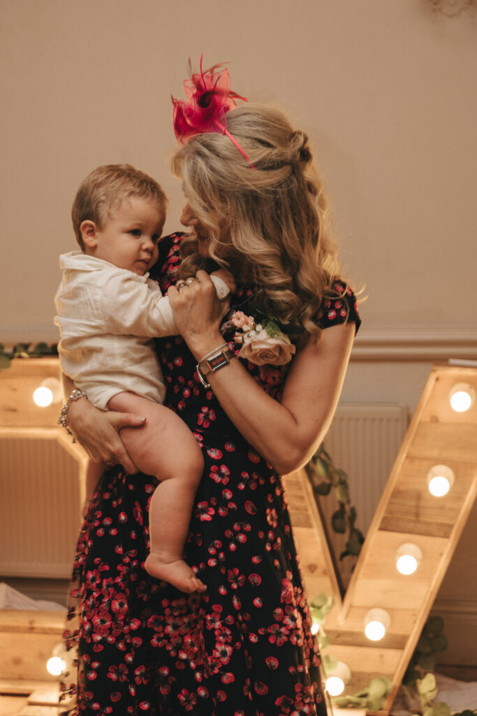 A woman with long blonde hair, wearing a black floral dress and a pink fascinator, holds a toddler in a white outfit. They stand in front of large illuminated wooden letters at Oaklands Hall. The setting appears festive, reminiscent of elegant wedding photography with decorative greenery and soft lighting. © Aimee Lince Photography