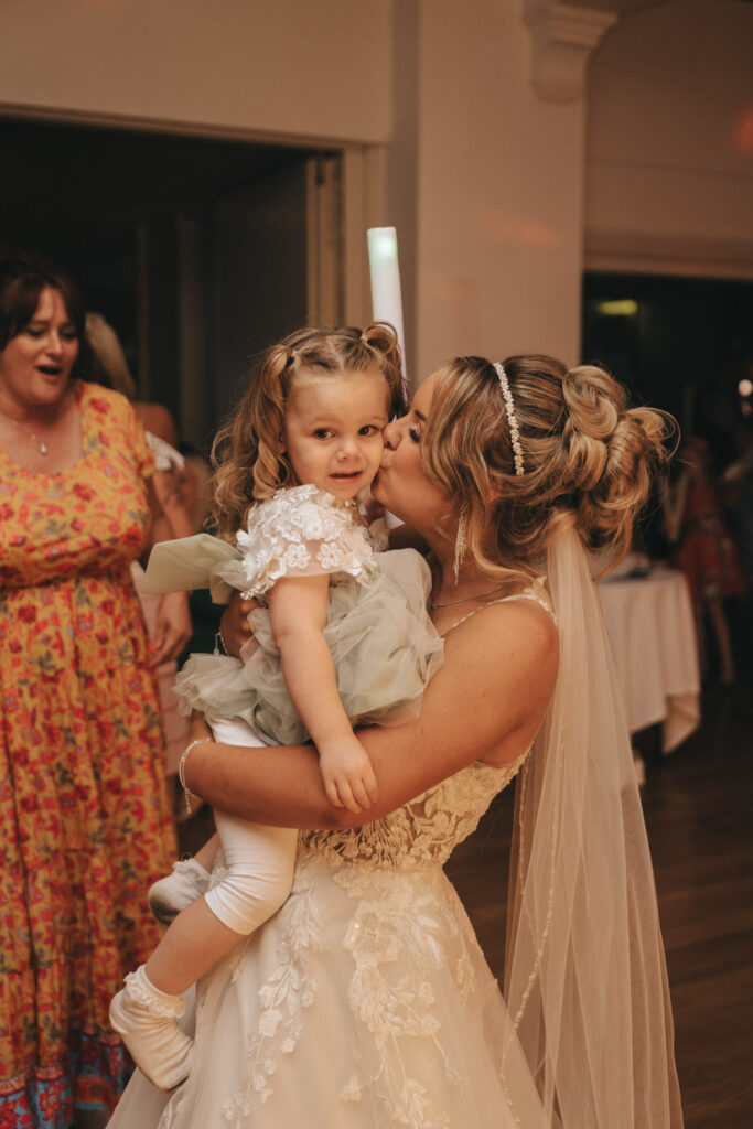 A bride in a white lace gown and veil holds a young girl with curly hair in a light blue dress, capturing a heartwarming moment of wedding photography. The bride kisses the child's cheek, both smiling, as a woman in a floral dress stands nearby at Oaklands Hall Hotel with its warm indoor lighting. © Aimee Lince Photography