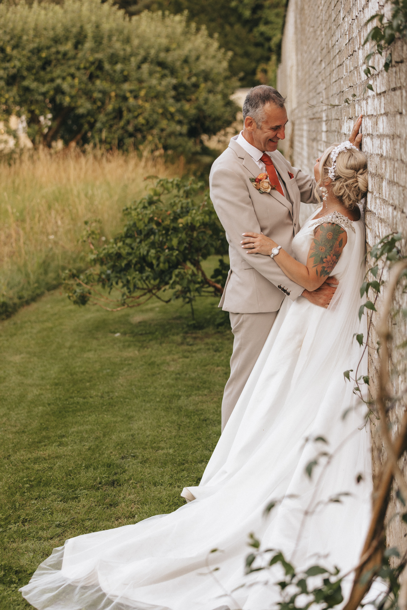 A groom in a beige suit and a bride in a flowing white gown stand outdoors against a stone wall, gazing at each other lovingly. The groom has one hand on the wall, leaning in, while the bride, adorned with tattoos, has her hands on his chest. Lush greenery surrounds them. © Aimee Lince Photography