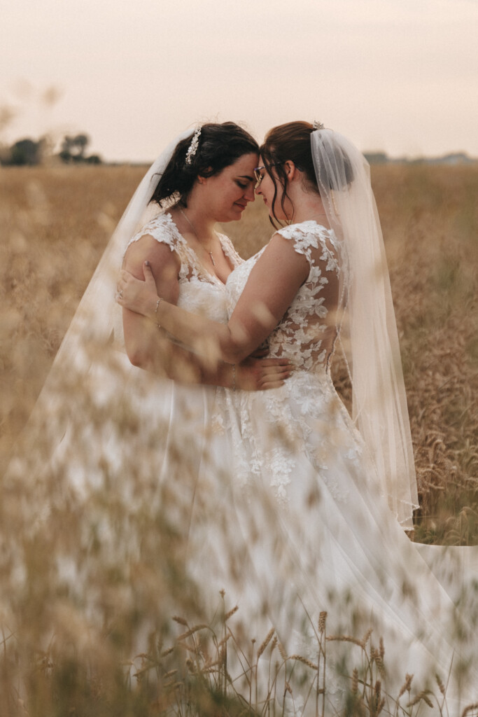 Two brides in white lace wedding dresses and veils embrace and share a tender moment in a field of tall grass. They stand nose to nose, smiling with eyes closed. The background features an open landscape with soft sunlight. Their joy and connection are palpable, creating a serene and intimate scene. © Aimee Lince Photography