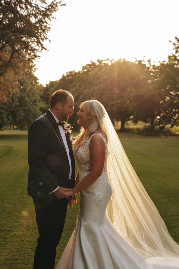 A bride and groom stand closely on a grassy lawn during sunset, holding hands and gazing at each other lovingly. The bride wears a white wedding dress with a long, flowing veil, while the groom is dressed in a black suit with a bow tie. Trees and sunlight are visible in the background. © Aimee Lince Photography