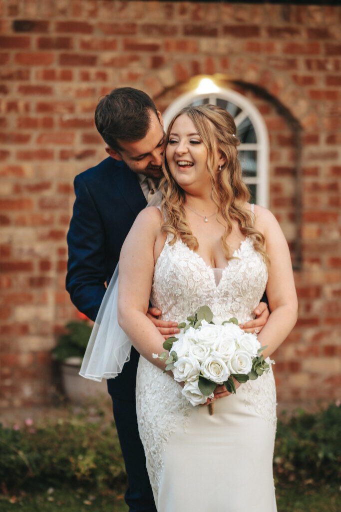 A couple poses in front of a brick wall with an arched window at Stallingborough Grange. The bride, wearing a lace wedding dress and holding a bouquet of white roses, smiles joyfully while the groom, in a navy suit, hugs her from behind and kisses her cheek. Both appear happy and in love. © Aimee Lince Photography