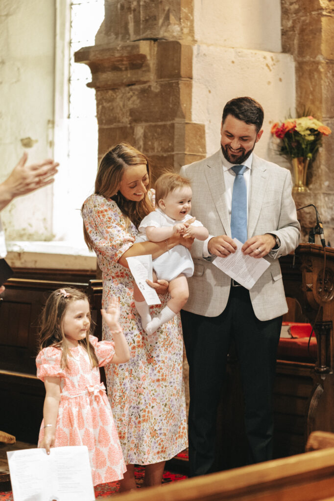 A woman holding a smiling baby, standing beside a man in a light-colored suit and tie, and a young girl in a peach dress. They are at All Saint's Church in New Waltham for Chester's Christening, near a stone wall and wooden pews. The woman and girl are holding papers. Flowers adorn a corner in the background. © Aimee Lince Photography