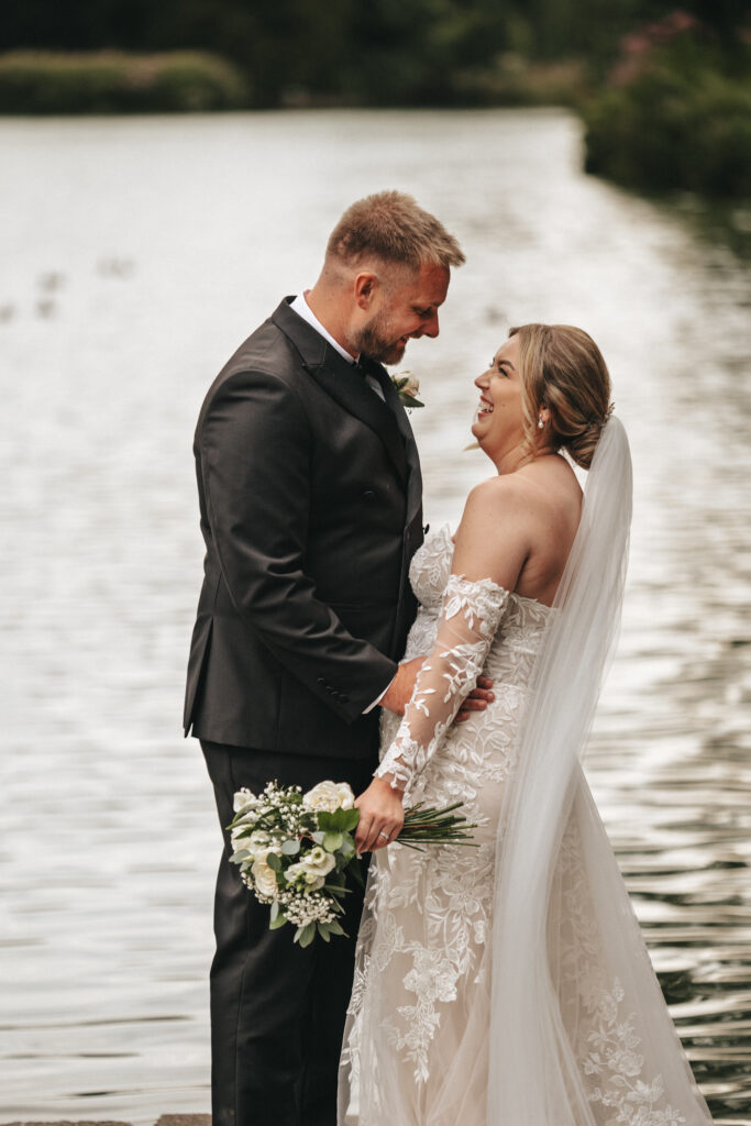 A bride and groom share a joyful moment by a serene body of water at Elsham Hall. The bride, in an off-shoulder lace gown and veil, holds a white flower bouquet. The groom is in a dark suit with a bow tie. Both are smiling at each other affectionately, standing close together with greenery in the background—a perfect wedding photography scene. © Aimee Lince Photography