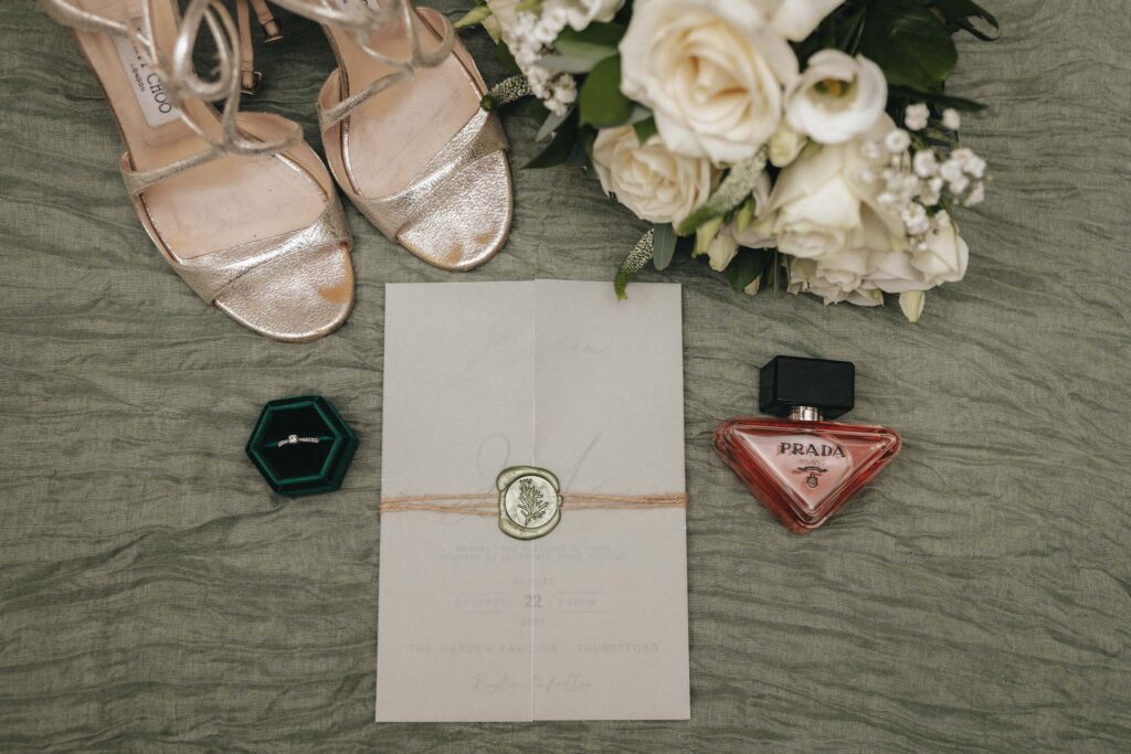 A stunning flat lay image captures the essence of elegance: gold sandals, a white flower bouquet, and a green jewelry box with a ring artfully arranged. A closed white envelope with a wax seal and a red Prada perfume bottle complete the tableau, set on textured green fabric—photography at its finest. © Aimee Lince Photography