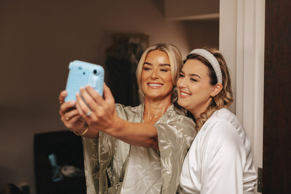 Two women are smiling and taking a selfie indoors, capturing the joy of the moment. One has blond hair and wears a light green robe, while the other with light brown hair and a headband dons a white robe. They stand close together, embodying fun wedding photography vibes against a beige wall background. © Aimee Lince Photography
