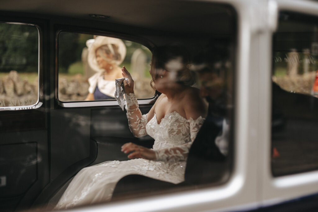 A bride in an off-shoulder lace dress is seated in the back of a car, waving. The window reflects light, partially obscuring her face. Outside, at Elsham Hall, a person in formal attire and a large hat stands blurred against a natural backdrop. The scene captures the essence of wedding photography. © Aimee Lince Photography