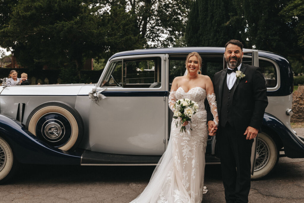 A bride in a lace wedding gown and a groom in a black tuxedo stand smiling, holding hands in front of a vintage car at Elsham Hall. The bride holds a bouquet of white flowers as trees and a child waving peek from behind—all captured perfectly by the wedding photographer. © Aimee Lince Photography