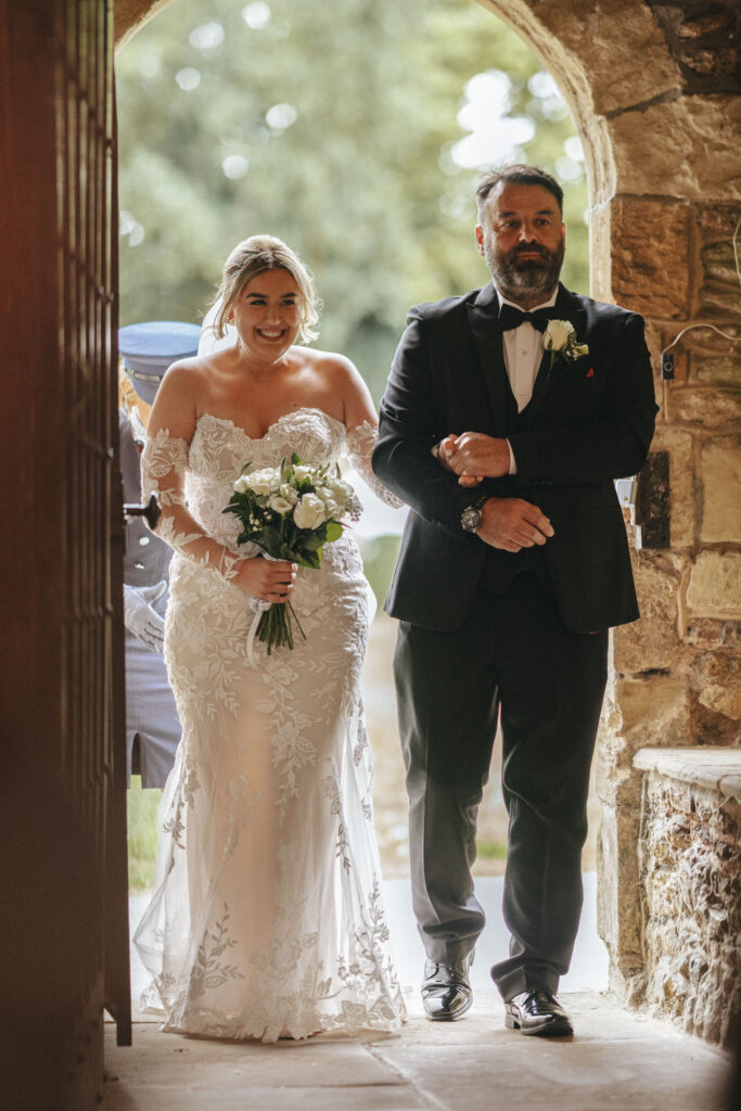 A bride in a white lace wedding dress and a man in a black suit with a bow tie enter through a stone archway at Elsham Hall. The bride, holding a bouquet of white flowers, smiles warmly as sunlight filters softly behind them, perfect for the wedding photographer to capture. © Aimee Lince Photography