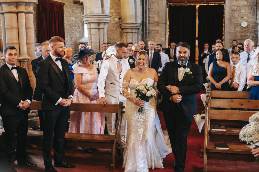 A bride in a lace gown holds a bouquet and walks down a church aisle with a man in a black suit, as the wedding photographer captures every smile from the guests standing on either side. A groom in a white suit waits at the front. The church features stone walls, wooden pews, and red carpeting. © Aimee Lince Photography