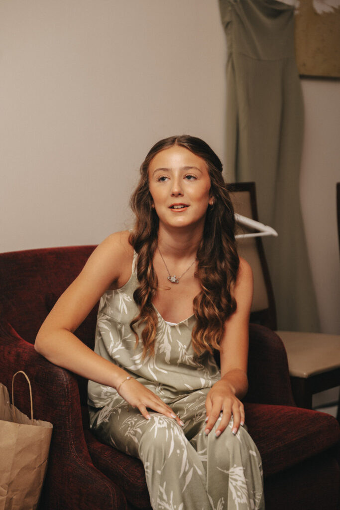 A young woman with long, wavy hair sits stylishly on a red armchair, showcasing an eye for photography. She wears a green sleeveless dress with a white floral pattern. A hanger with a green garment lingers behind her, while a paper bag rests to her left in this elegant indoor setting. © Aimee Lince Photography