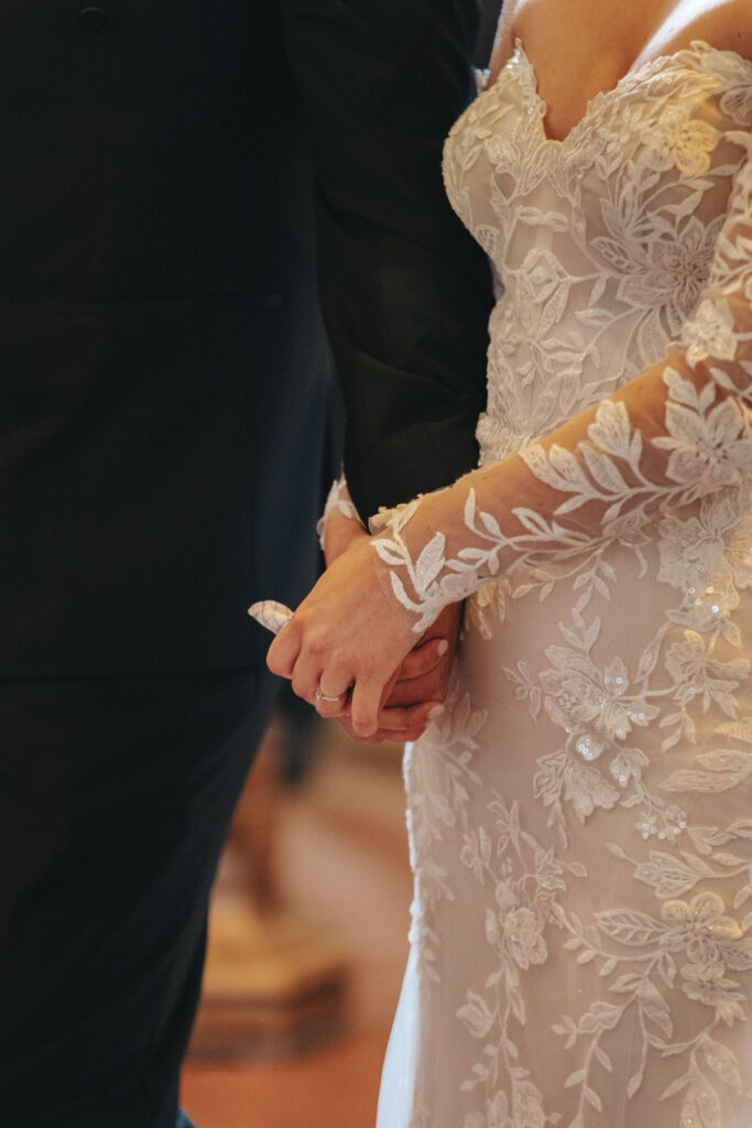 A bride and groom hold hands at Elsham Hall. The bride wears a white lace wedding dress with floral patterns and sheer sleeves, while the groom is in a black suit. The photographer captures this intimate moment with a soft blur in the background, emphasizing their hands and attire during the ceremony. © Aimee Lince Photography
