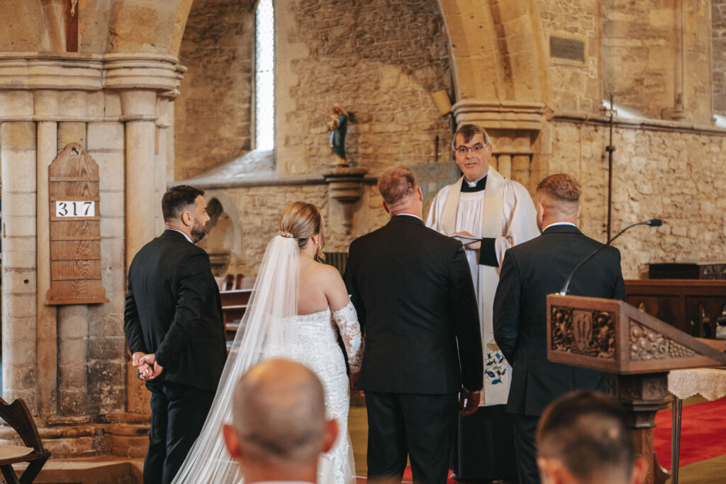 A wedding photographer captures a beautiful ceremony inside a stone church. A bride in a white dress with a long veil stands beside three men in black suits facing the priest. The priest, in a white robe, holds a book and speaks to the couple, all framed by arched stone architecture and stained glass windows. © Aimee Lince Photography