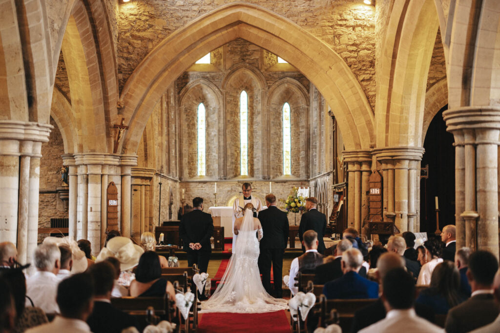 A wedding photographer captures the enchanting ceremony in a stone church with high arches and stained glass. At the altar, the bride and groom exchange vows while guests watch from pews. The warmly lit interior of Elsham Hall features floral arrangements that enhance the romantic setting. © Aimee Lince Photography