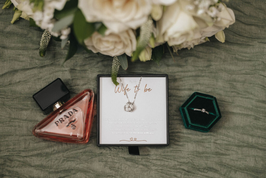 A triangular Prada perfume bottle, a diamond engagement ring in a green hexagonal box, and a "Wife to be" card with earrings are artfully arranged on green fabric. White flowers peek from the top, creating an enchanting scene perfect for wedding photography at Elsham Hall. © Aimee Lince Photography