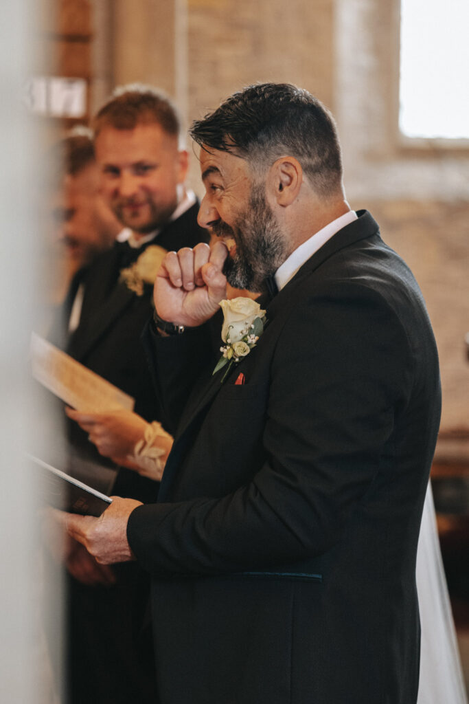 A man in a black suit with a boutonniere laughs, holding a piece of paper, as sunlight streams through the window at Elsham Hall. Nearby, another man in a similar suit smiles behind him. This moment, captured by the photographer's lens, suggests they're at an elegant formal event. © Aimee Lince Photography