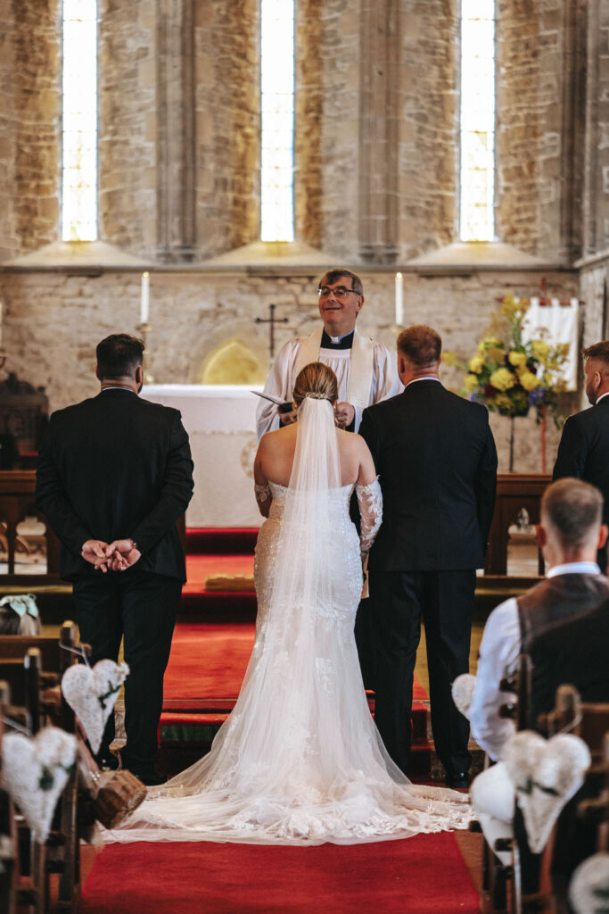 A bride in a white lace gown and veil stands facing an officiant in a stone church with large windows. Two men in suits stand beside her. Capturing this moment, a wedding photographer discreetly moves among the guests seated with aisle decorations. The altar glows with natural light and floral arrangements. © Aimee Lince Photography