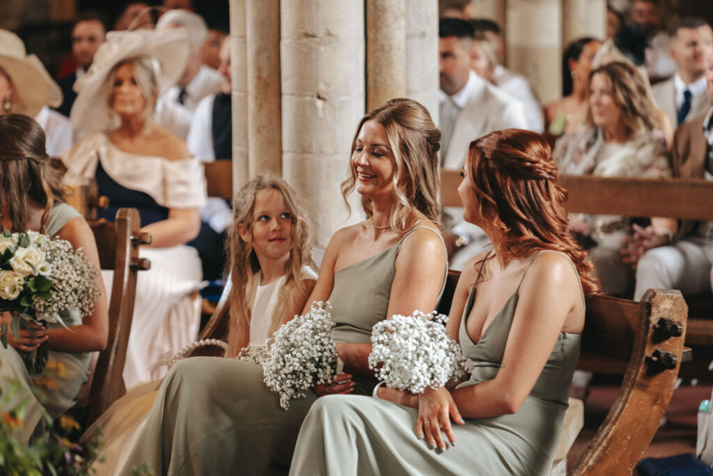 Three women in sage green dresses and a young girl in white sit on wooden pews at Elsham Hall, smiling with bouquets of white baby's breath. Behind them, guests in formal attire fill the church. The setting is warm and intimate, with soft natural light highlighting the wedding scene. © Aimee Lince Photography