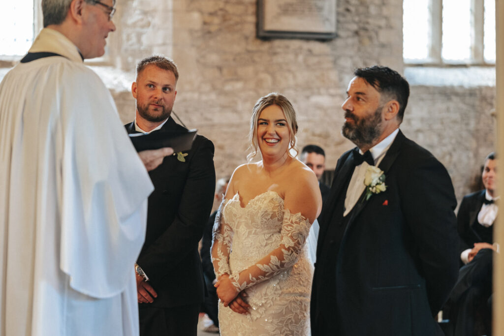 A bride in a white lace dress stands smiling between two men in suits during the wedding ceremony in a stone church. One man is bearded, and the other is clean-shaven. A clergyman in a white robe addresses them with seated guests visible, as the photographer captures this cherished moment. © Aimee Lince Photography