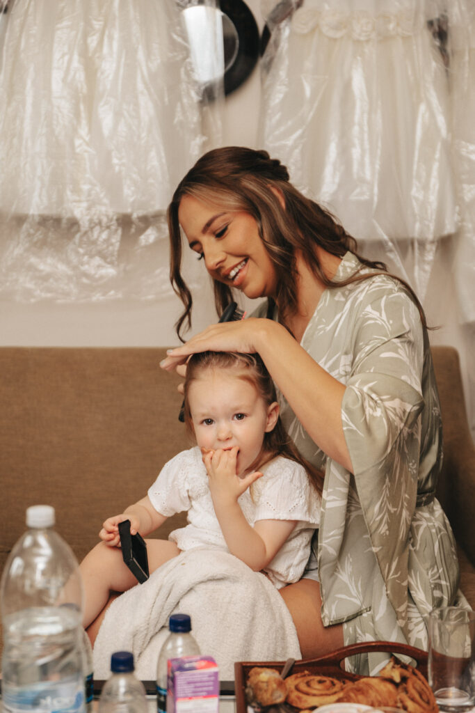 A woman in a floral robe sits on a couch, smiling as she styles a toddler's hair for wedding photography. The toddler, wearing a white dress, holds a comb while seated on a towel. Nearby, water bottles and a snack tray rest on the table, and garments hang in the background. © Aimee Lince Photography