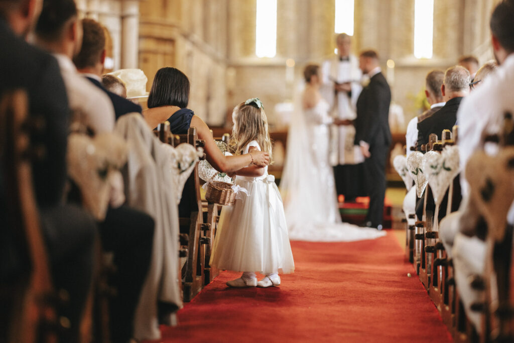 In the serene setting of Elsham Hall, a young flower girl in a white dress and floral headband clutches her basket as she stands on the red carpeted aisle. Guests seated on ornate chairs with decorative bows watch, while wedding photography captures the couple and officiant at the altar, softly out of focus. © Aimee Lince Photography
