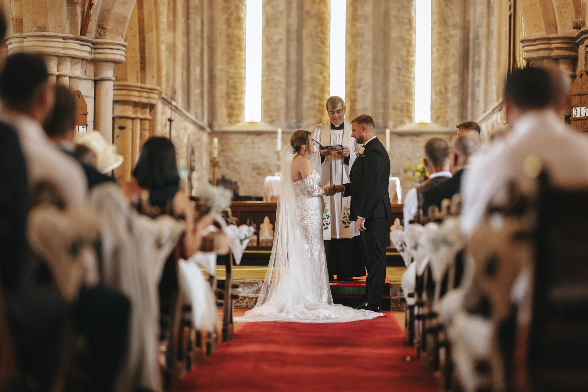 Elsham Hall Wedding Photographer - Gabriella & Alex © Aimee Lince Photography (237) A bride and groom stand at the altar in a church, facing each other. The wedding photographer captures every moment as the priest reads from a book. Guests sit on either side of the aisle on chairs adorned with white decorations. Tall stone columns and large windows let in natural light, illuminating the scene. © Aimee Lince Photography