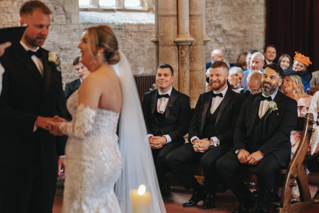 In the stone-walled ceremony room at Elsham Hall, a bride in a white gown and lace sleeves stands facing her groom, who wears a black suit with a white rose boutonniere. A wedding photographer captures the moment as guests, including three suited men in the front row, watch intently. © Aimee Lince Photography