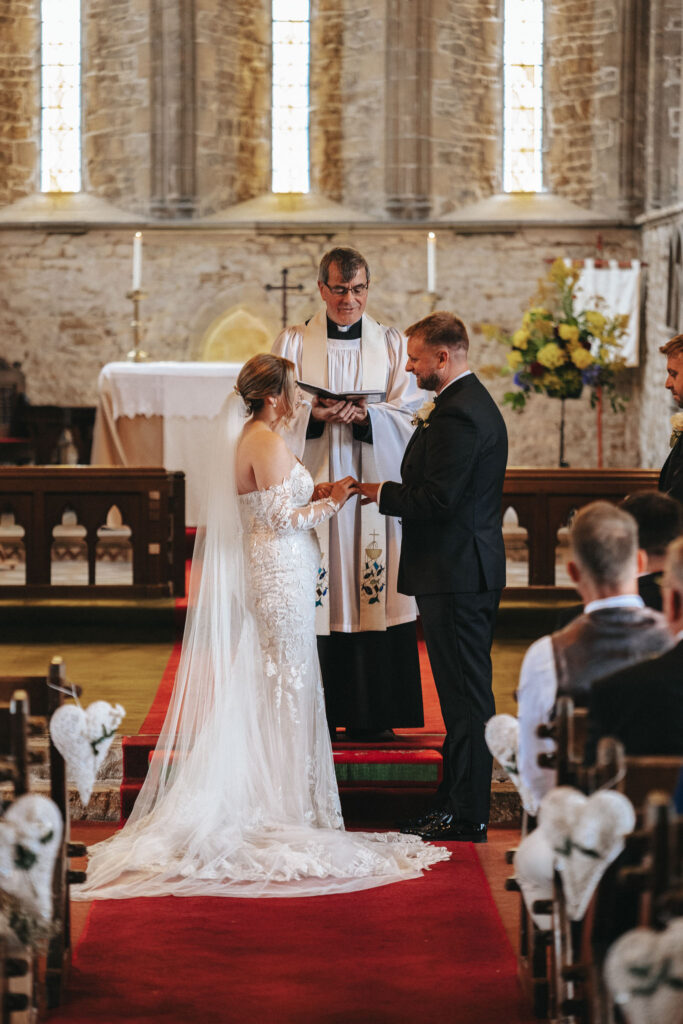 A bride in a white, lace, off-the-shoulder gown with a long veil stands facing the groom, who is in a black suit. They hold hands in front of the clergyman at Elsham Hall. A wedding photographer captures the moment amidst flowers and candles, while guests sit beside an aisle adorned with white bows.
. © Aimee Lince Photography