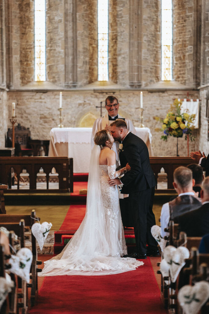 In a beautifully captured moment by the wedding photographer, a bride and groom share a kiss at Elsham Hall. The bride's off-shoulder lace gown and the groom's black suit complement the altar's floral elegance, as guests seated along the bow-adorned aisle witness their joy. © Aimee Lince Photography