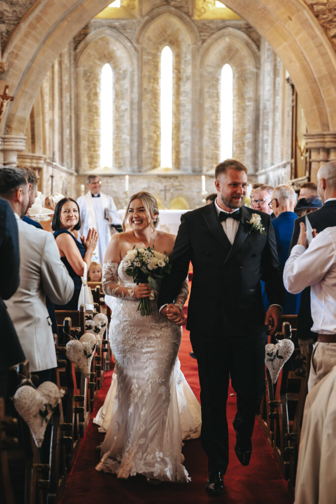 A bride and groom smile as they walk hand-in-hand down the aisle of a church, captured beautifully by the wedding photographer. The bride wears a white lace gown and holds a bouquet, while the groom is in a black tuxedo. Guests stand and clap amidst stone arches and tall, narrow windows. © Aimee Lince Photography