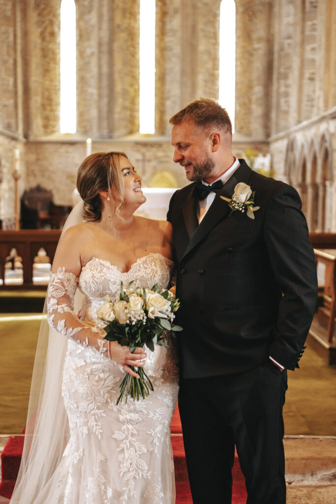 A bride and groom stand together inside a church, captured in stunning photography. The bride wears a lace wedding gown, holding a bouquet of white roses, while the groom is in a black tuxedo with a white boutonnière. They gaze at each other affectionately amid the stone interior and stained glass windows. © Aimee Lince Photography
