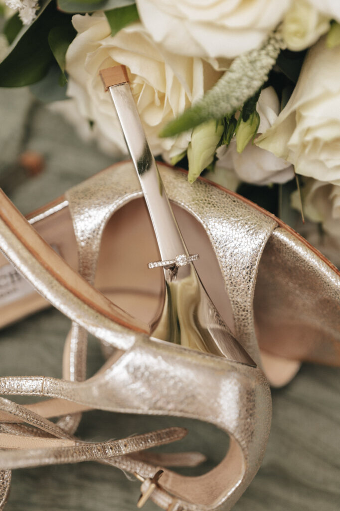 A sparkling engagement ring rests on the heel of a metallic high-heeled shoe. The shoe is silver with a textured surface. Behind it, white roses and greenery add a soft, elegant backdrop, all set on muted green fabric—an exquisite setting captured by the keen eye of a wedding photographer. © Aimee Lince Photography