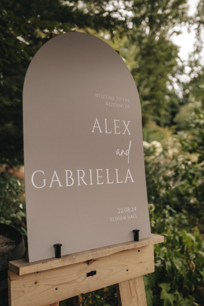 A taupe wedding sign reads "Welcome to the Wedding of Alex and Gabriella" in elegant, white lettering. The date "22.08.24" and location "Elsham Hall" grace the bottom, perfect subjects for any wedding photographer. Mounted on a wooden stand, it stands amid a lush, green garden backdrop. © Aimee Lince Photography